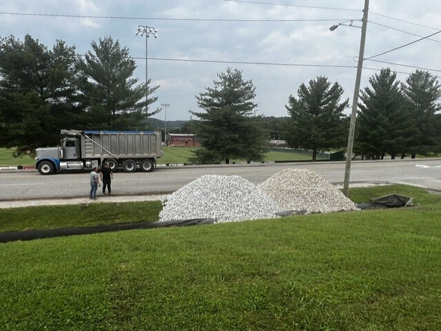 A large truck is parked on the side of the road. Two people stand nearby. Piles of gravel are on the grass.