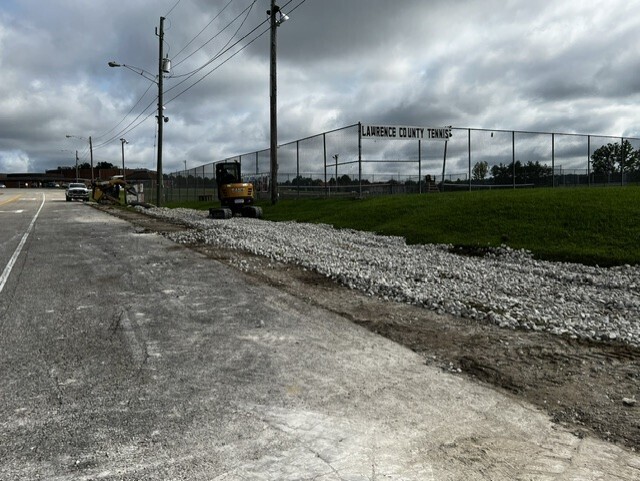 An empty road with gravel on the side, a yellow machinery vehicle parked, a fence, and a cloudy sky.