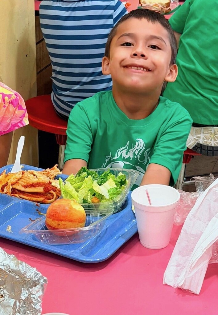 A young boy in a green shirt sitting at a cafeteria table with a meal tray in front.