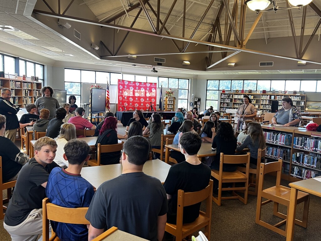 A group of people sits around tables in a library. Some are facing forward, others sideways. A large red banner hangs in the background.