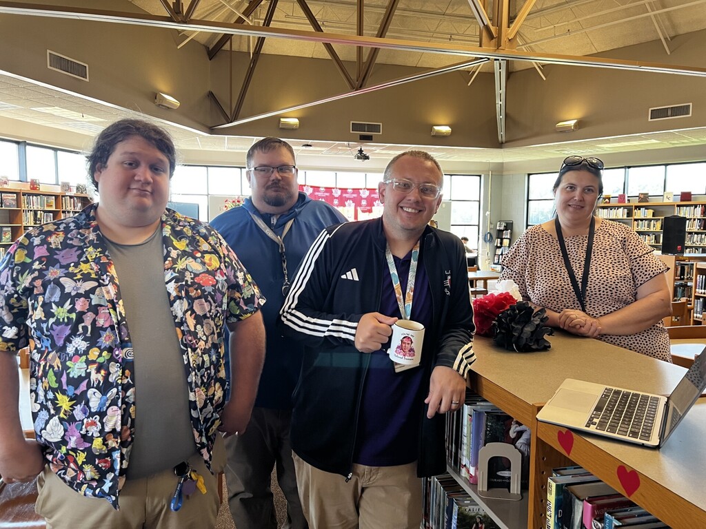 Five people stand near a desk with a laptop in a library. One holds a mug.