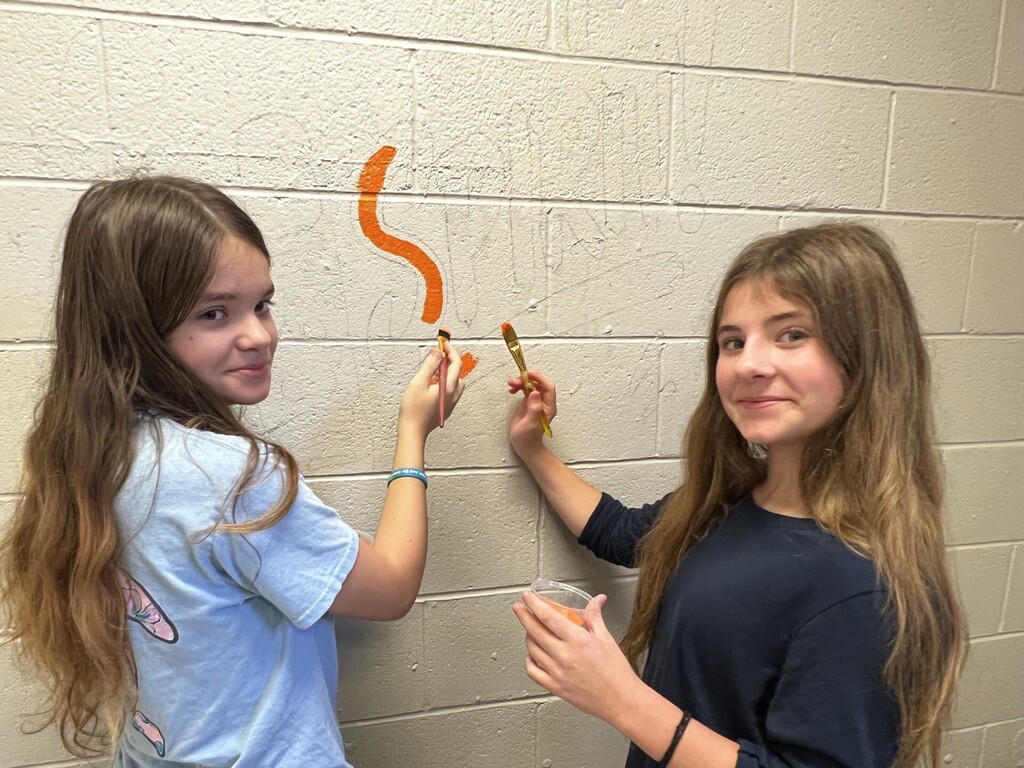 Two young girls paint an orange, wavy line on a white wall. One holds a paintbrush, and the other holds a paint container.