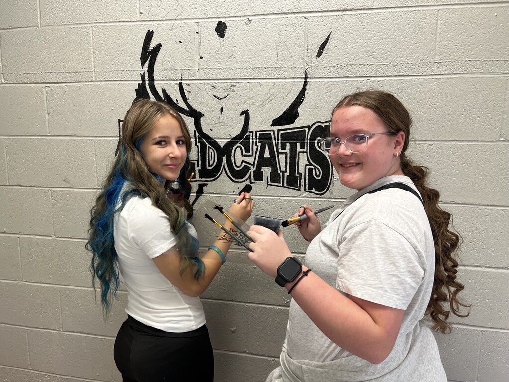 Two women, one with blue hair and the other with glasses, painting a wall with the word "Dcats".