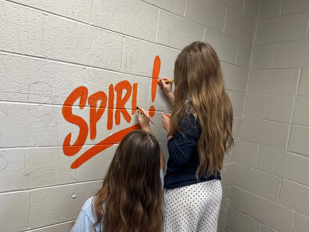 Two women painting the word "SPIRIT" in orange on a white wall in a corner.