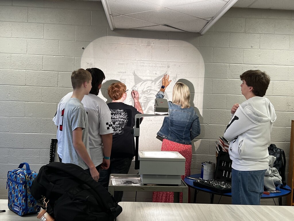 A group of people, possibly students, stand around a large whiteboard with a cat drawing. They are indoors.