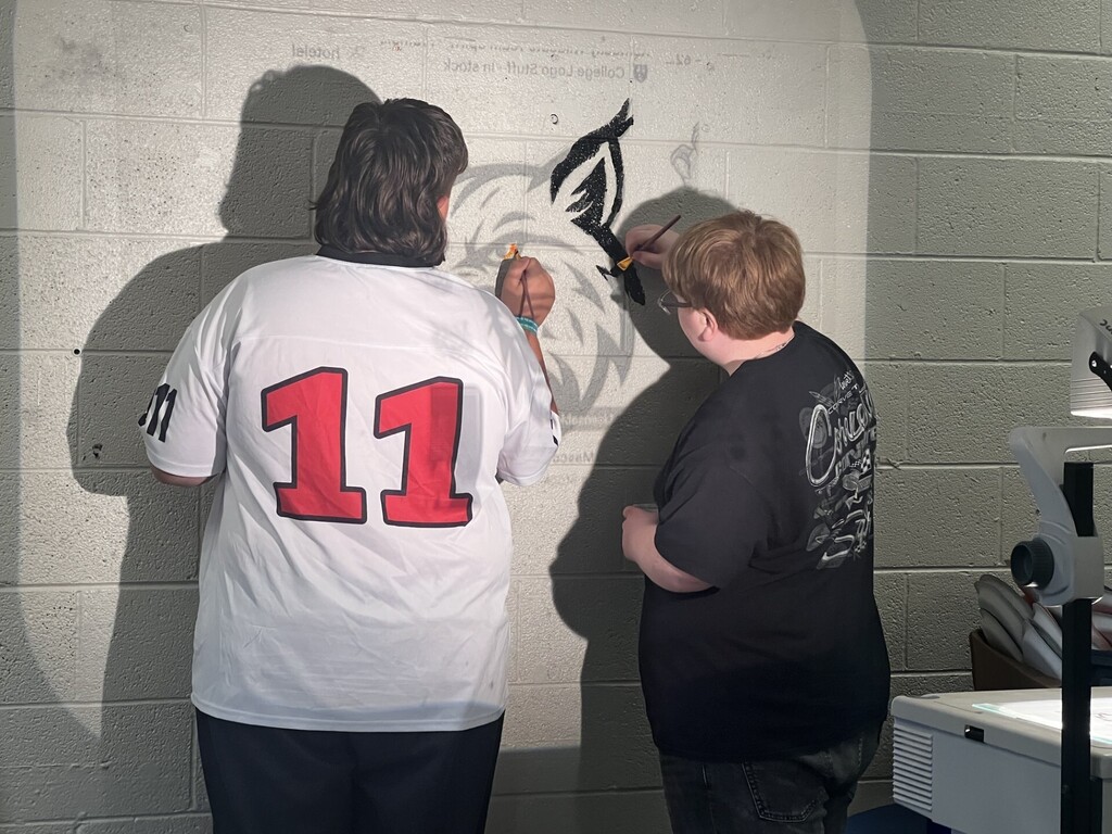 Two people standing next to a wall, one painting a wolf design, the other holding a paintbrush.