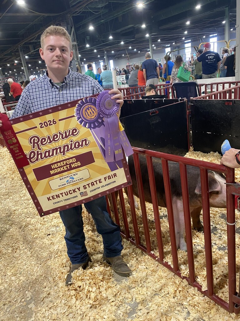 A man with a Reserve Champion sign stands in an indoor livestock pen, holding a blue ribbon.