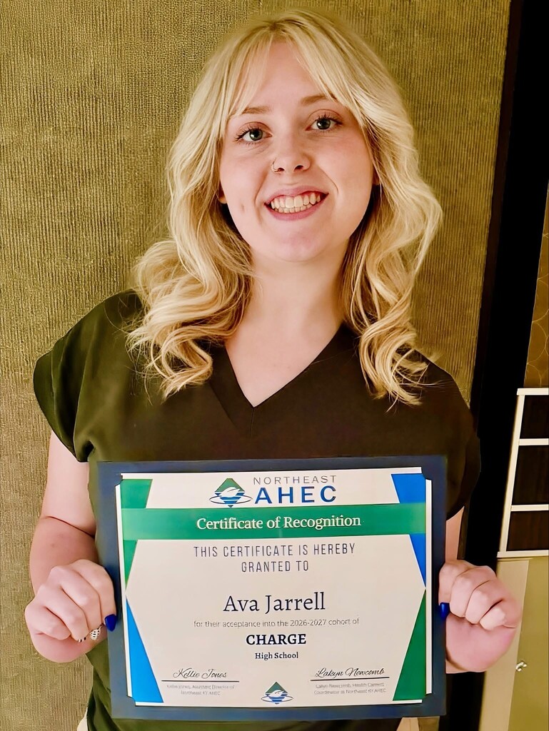 A young person with blonde hair holds a framed certificate with a green background. The certificate reads "Ava Jarrell."