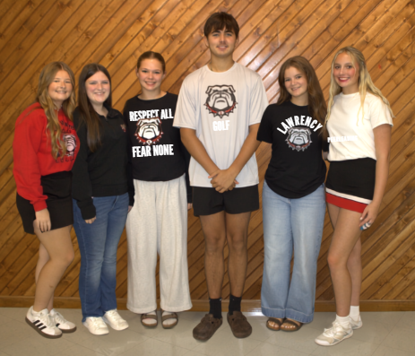 Five young adults stand together, smiling for a photo. Three wear white, and two wear red. Behind them, a wooden wall.