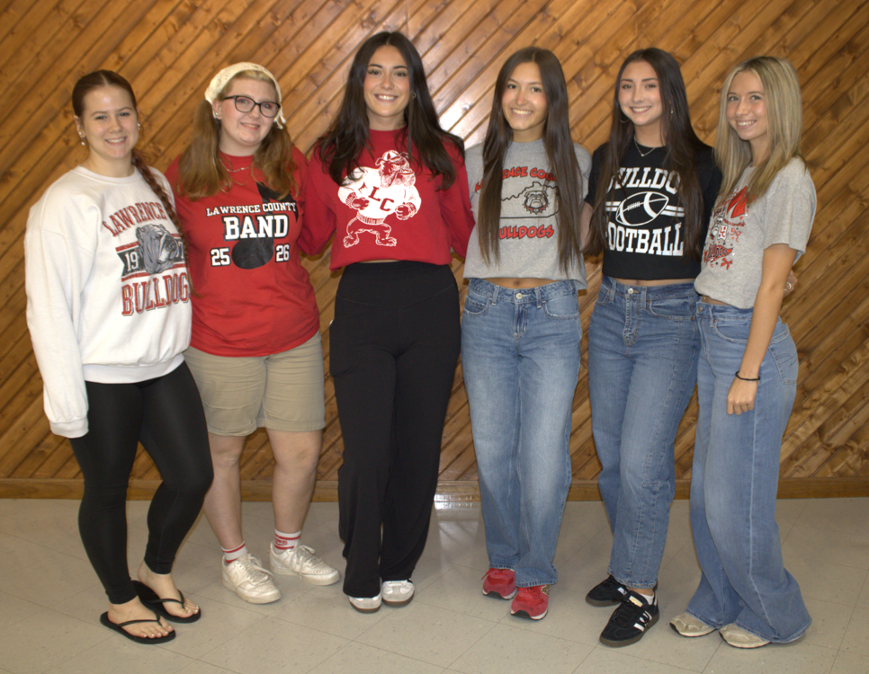 Five young women in matching tops and different bottoms stand in a row against a wooden wall.