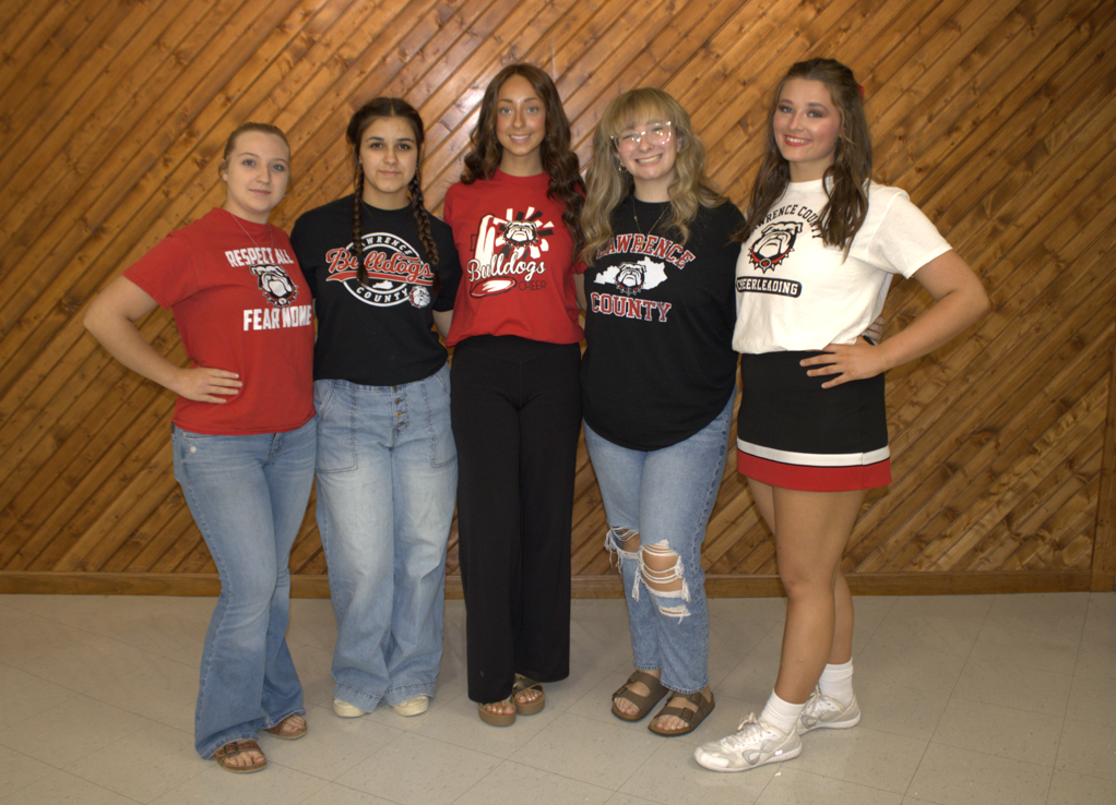 Five young women stand together against a wooden wall. They wear varied school-themed attire, including red and black shirts and a white t-shirt.