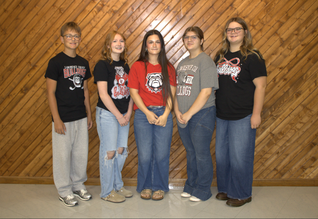 Five people in casual attire, with different colored tops and jeans, standing in front of a wooden wall.