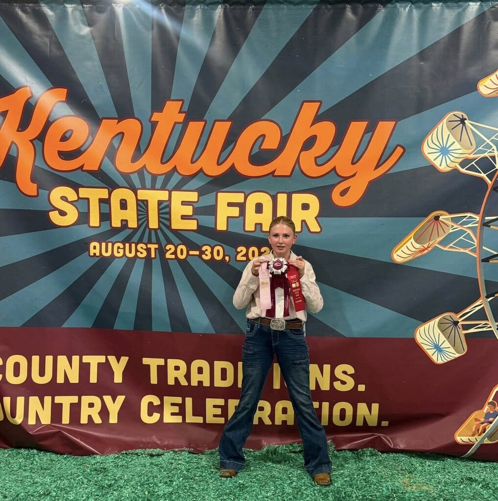 A young person stands before a banner for the Kentucky State Fair, holding a medal.