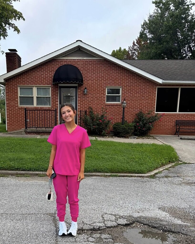 A woman in pink scrubs stands in front of a brick house with a green lawn.