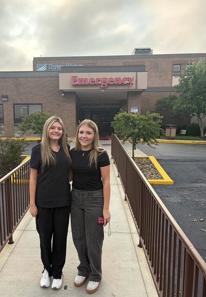 Two women in black uniforms stand on a hospital's sidewalk, with trees and a fence in the background.
