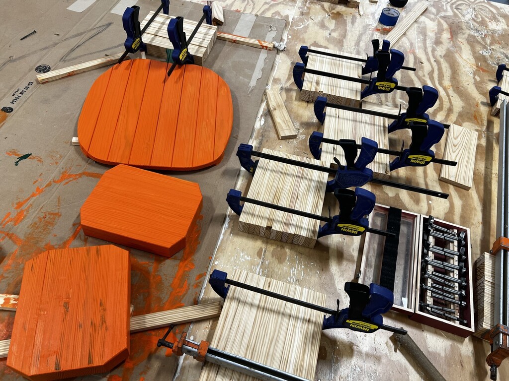 A workshop table displays four orange wooden boards secured by blue clamps. A tool box and assorted tools are nearby.
