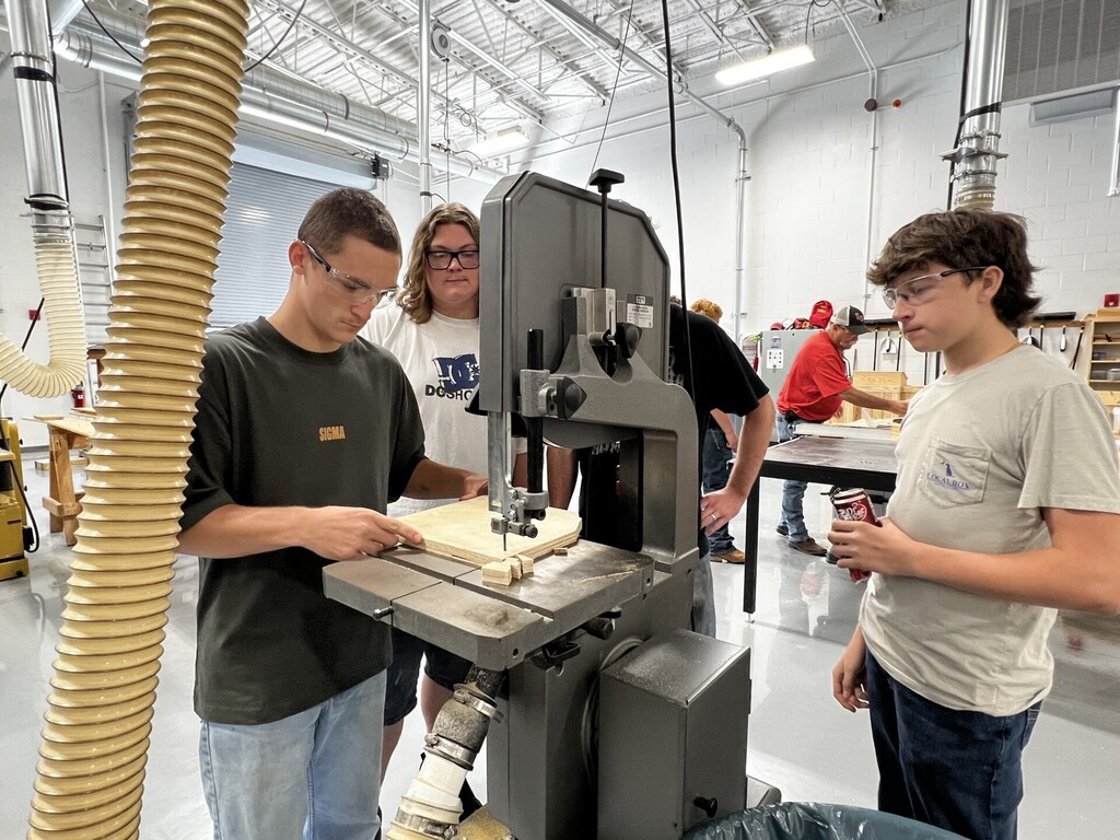 Several people observe a man operating a large machine in a wood shop. One man leans over to inspect the machine.