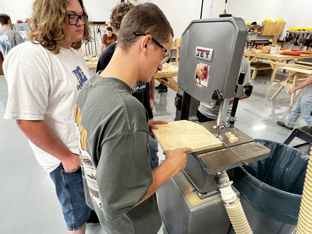 Two men are in a workshop. One is cutting wood on a saw while the other watches.
