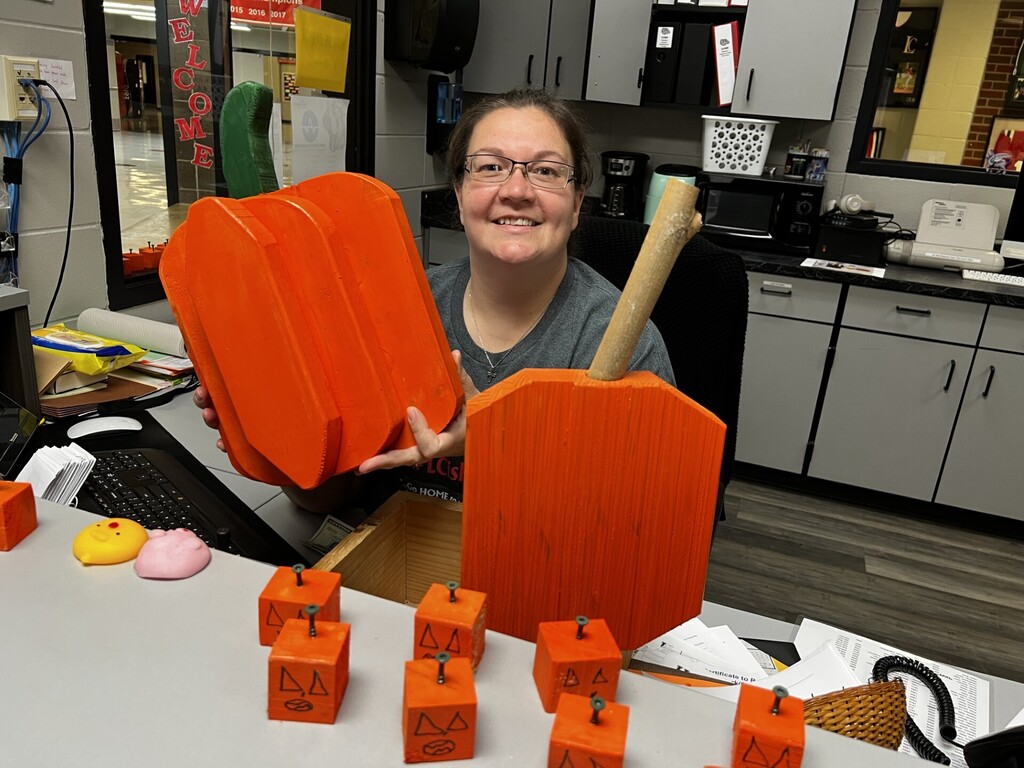 A woman with glasses holding three orange pumpkins sits at a desk with eight smaller orange pumpkin-shaped objects.