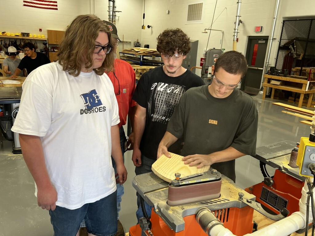 Four individuals in a workshop, one bent over a machine, another holding a piece of paper.