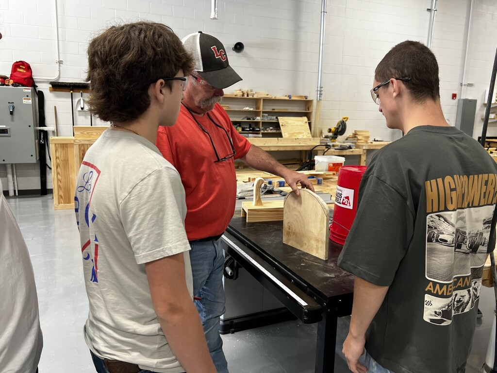 Three people are in a workshop, looking at a wooden structure on a table. A man in a red shirt is explaining.