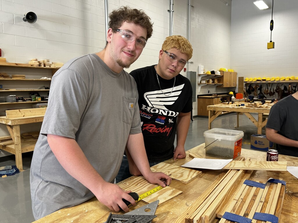 Two young men are working together in a workshop, one wearing glasses and measuring a wooden plank.