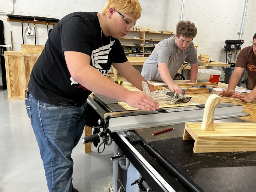 Three people in a workshop. One in a black shirt is cutting wood with a saw.