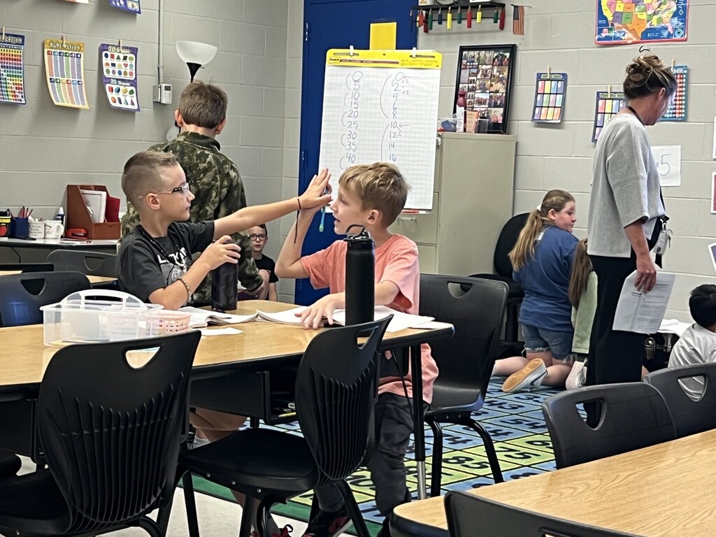 Classroom with students sitting at tables. One student has hand on forehead while another holds up a pencil.