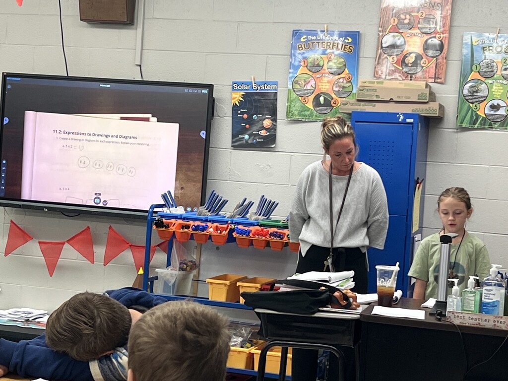 A teacher stands at the front of a classroom. Students sit in front, watching a presentation on a screen.