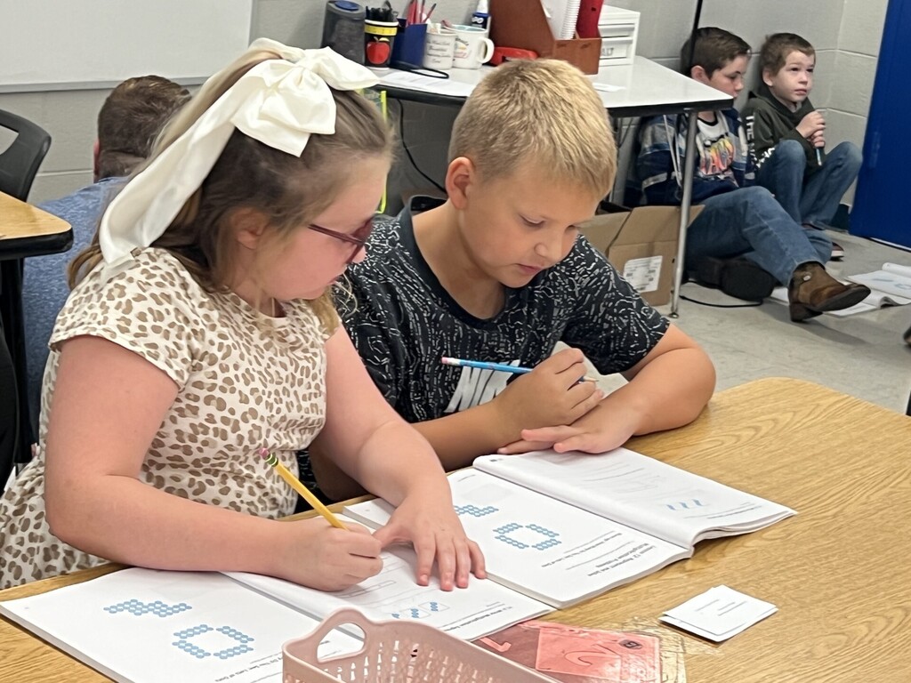 Two children, one with glasses and a bow, the other with brown hair, are writing in a book.