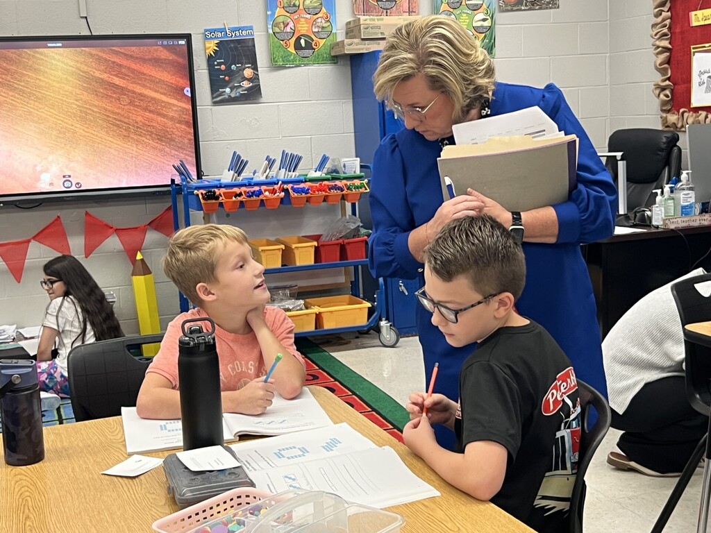 A woman in a blue robe assists two students at a desk. One student looks up, and the other writes.