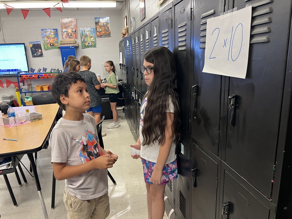 Two children, a boy and a girl, are in a classroom, standing by lockers. The girl has long hair.
