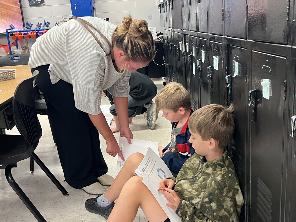 An adult woman holds a paper and guides two children sitting by lockers, reading papers, in a classroom.