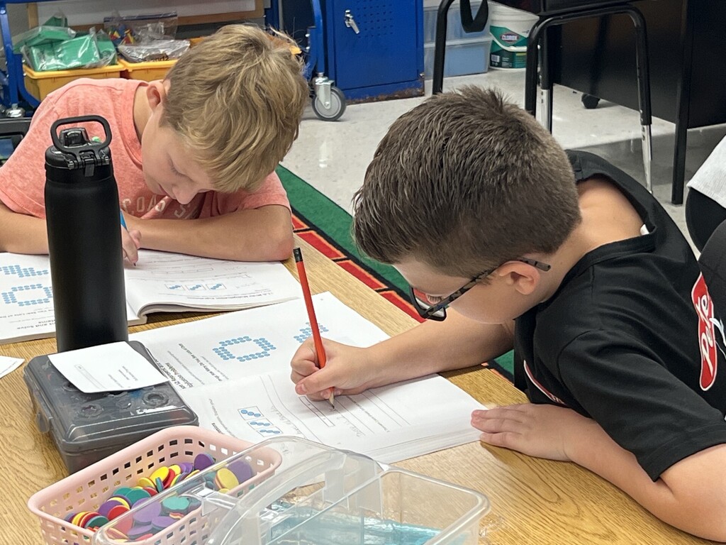 Two students seated at a desk in a classroom. One is writing with a pencil, another looks over.