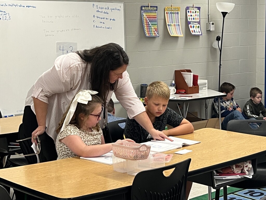 A woman helps two children at a desk. A white board with papers is on the wall.