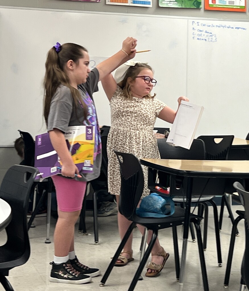 Two young girls, one with glasses, stand in a classroom. One holds a book, the other holds a paper, and they are pointing. Chairs and desks are around them.