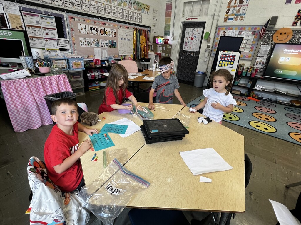 Four children sit at a table in a classroom, focused on their work. A table holds various items. Brightly colored decor is on the walls.
