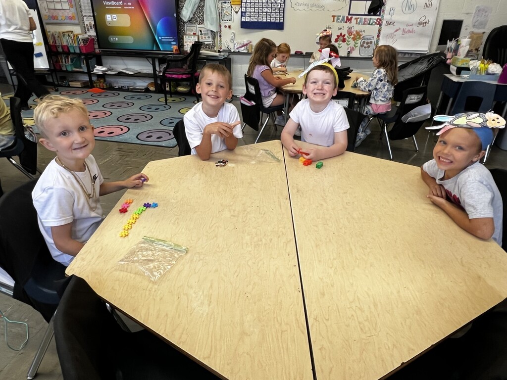 Four children in a classroom sit at a large wooden table, smiling and playing with objects.
