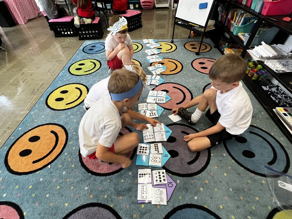 Four kids are playing with cards on a rug with smiley faces. One child is wearing a headband.