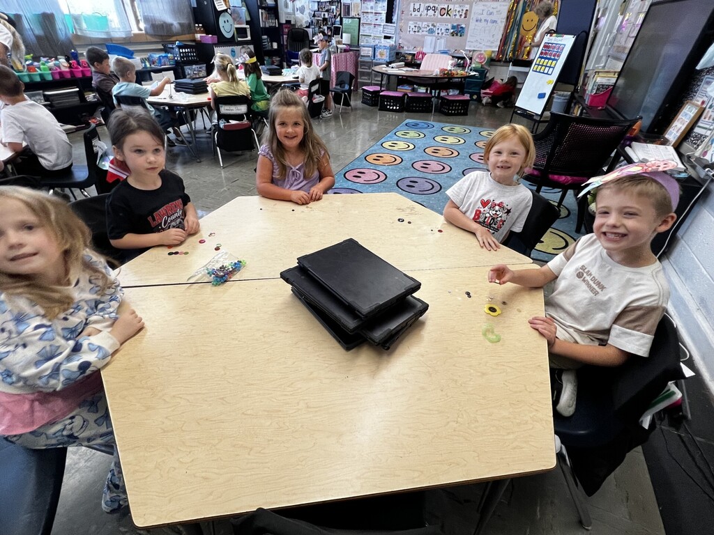 Five children sit around a table in a classroom, engaging in an activity. A carpeted floor is visible, and various classroom items are in the background.