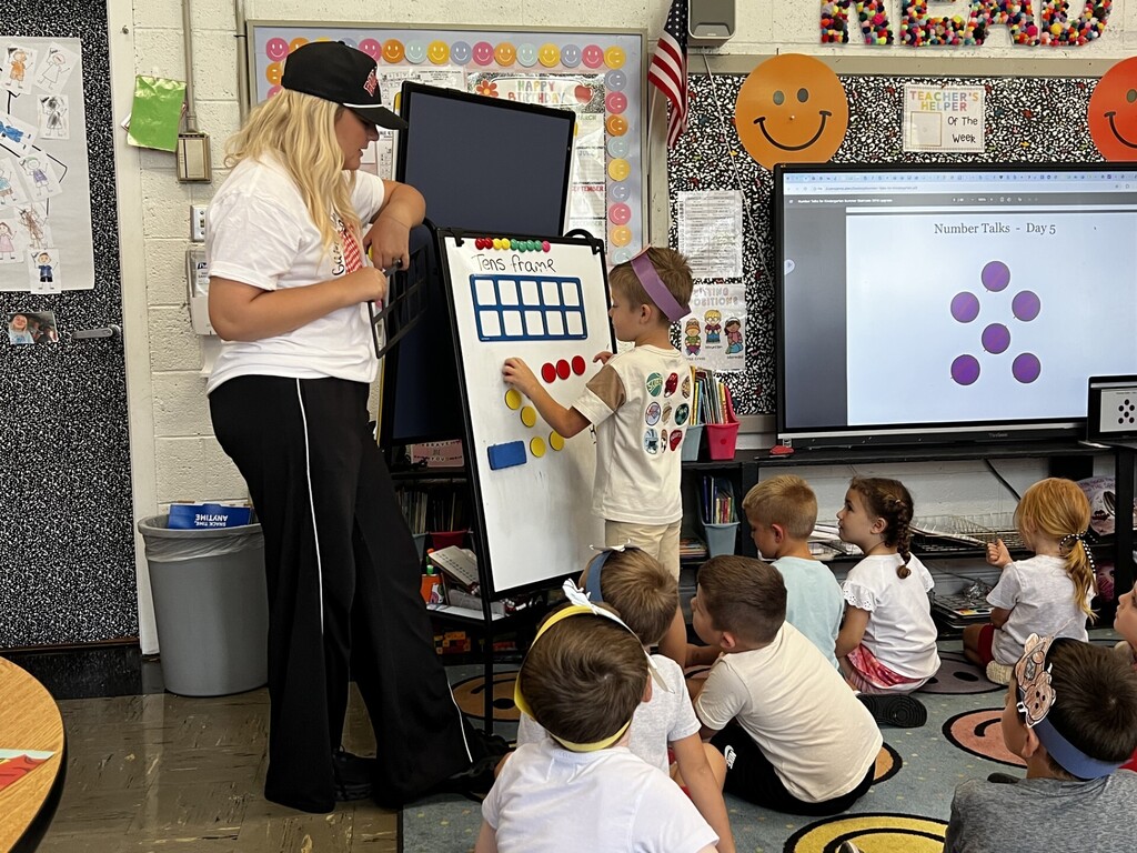 Woman in hat and cap teaches young children using a board while they sit in a classroom.