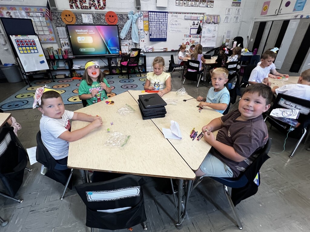 Several children sit at tables in a classroom. They engage in activities while a monitor displays information in the background.