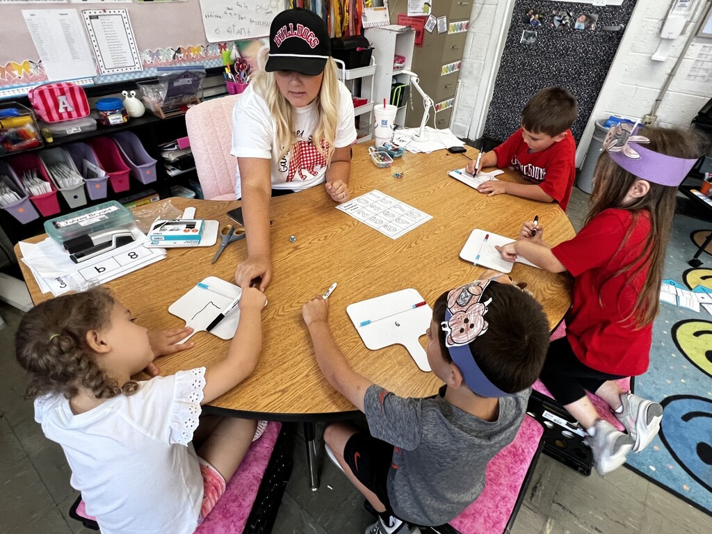 A teacher helps four children at a table, all wearing headbands. They use writing utensils on paper.