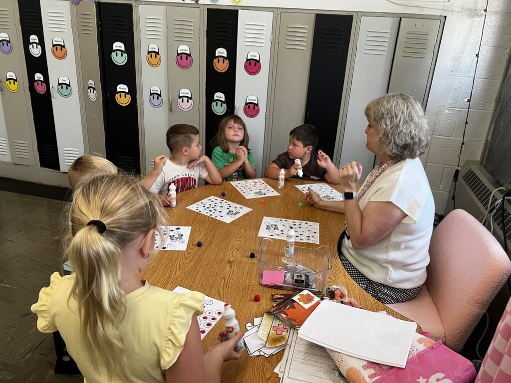 A group of children and an adult sitting around a table in a classroom. The table is covered with art supplies.