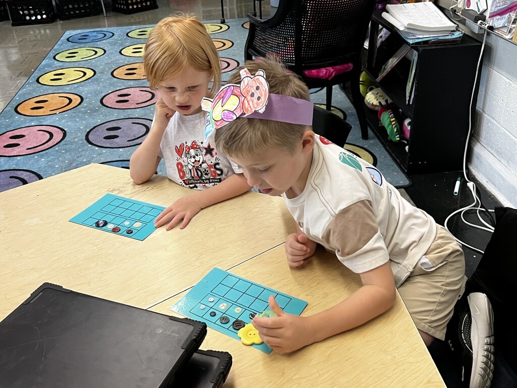 Two children seated at a table, engaged with a blue game board and cards. Background features a colorful rug and a desk with books and a chair.