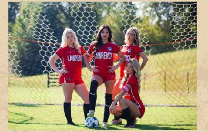 Five female soccer players in red uniforms posing in front of a goal on a grassy field.