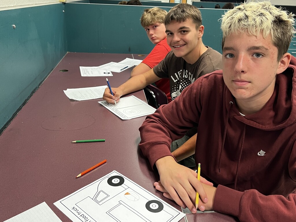Three boys at a table with papers and pencils. One holds a pen and looks at the camera.