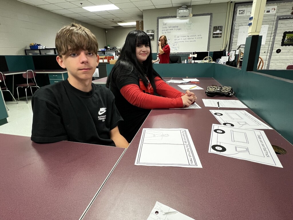 Two people sit at a table with papers in a room with a whiteboard and other people.