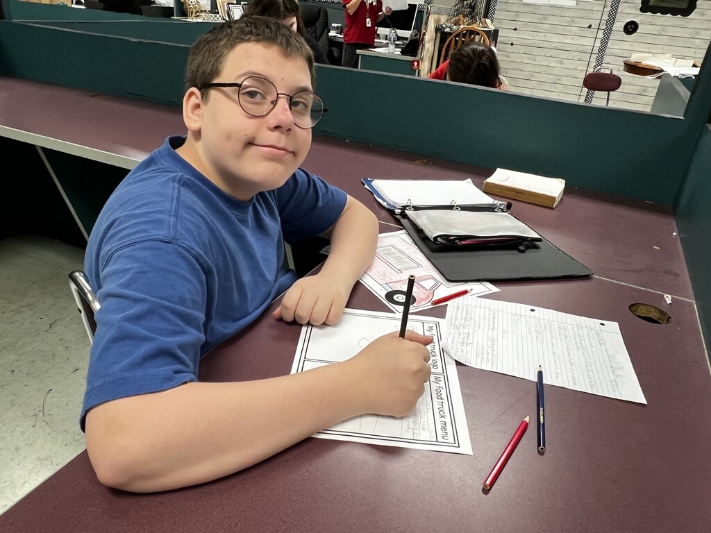 Person in blue shirt seated at a desk, writing on paper with several pens and papers around.