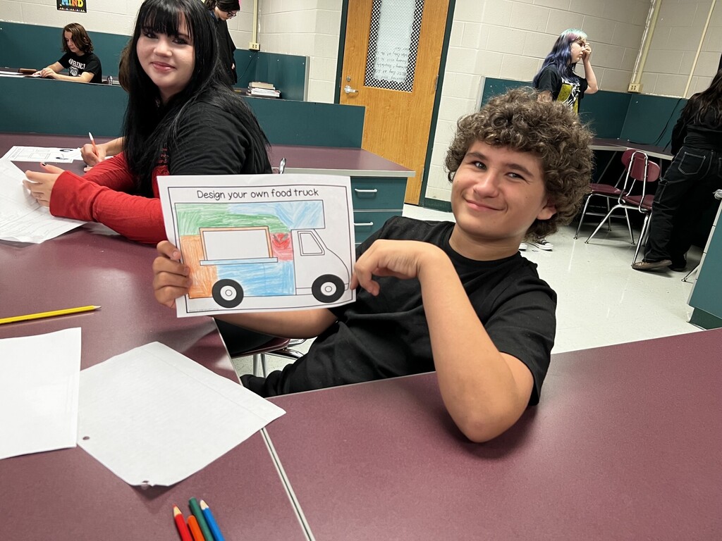 A young boy displays a coloring page of a food truck while sitting at a desk.